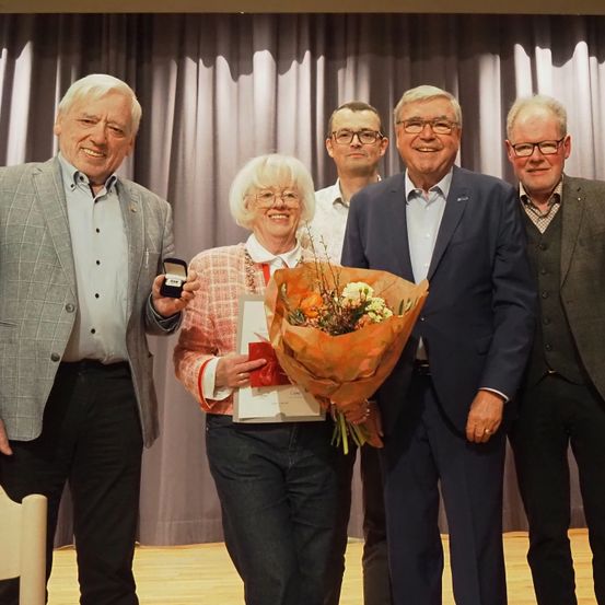 Fünf Erwachsene, darunter eine Frau, die Blumen und ein Zertifikat hält, stehen hinter einem Vorhang für ein Foto. Die Frau lächelt und hält einen Ring. Der Mann links von ihr hat einen Ring in der Hand.