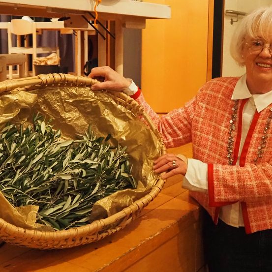 Eine ältere Frau mit Brille lächelt und hält einen Korb mit goldumwickelten Blättern. Sie trägt eine rote karierte Jacke mit einer Halskette. Hinter ihr ist eine gelbe Wand mit einem Metallgriff.