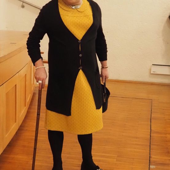 Eine Frau in einem gelben Kleid und schwarzem Cardigan hält einen Stock. Sie trägt eine Halskette, Armband und schwarze Strümpfe. Sie steht auf einem Holzboden in der Nähe einer weißen Wand.