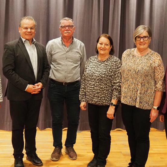 Fünf Personen stehen nebeneinander vor einem Vorhang und lächeln für ein Foto. Drei Männer und zwei Frauen sind in Freizeitkleidung gekleidet.