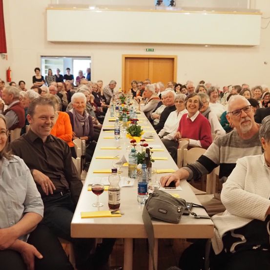 Eine große Gruppe von Menschen sitzt an einem langen Tisch mit Blumen, Flaschen und Gläsern. Der Tisch ist für eine Mahlzeit oder Zusammenkunft gedeckt.