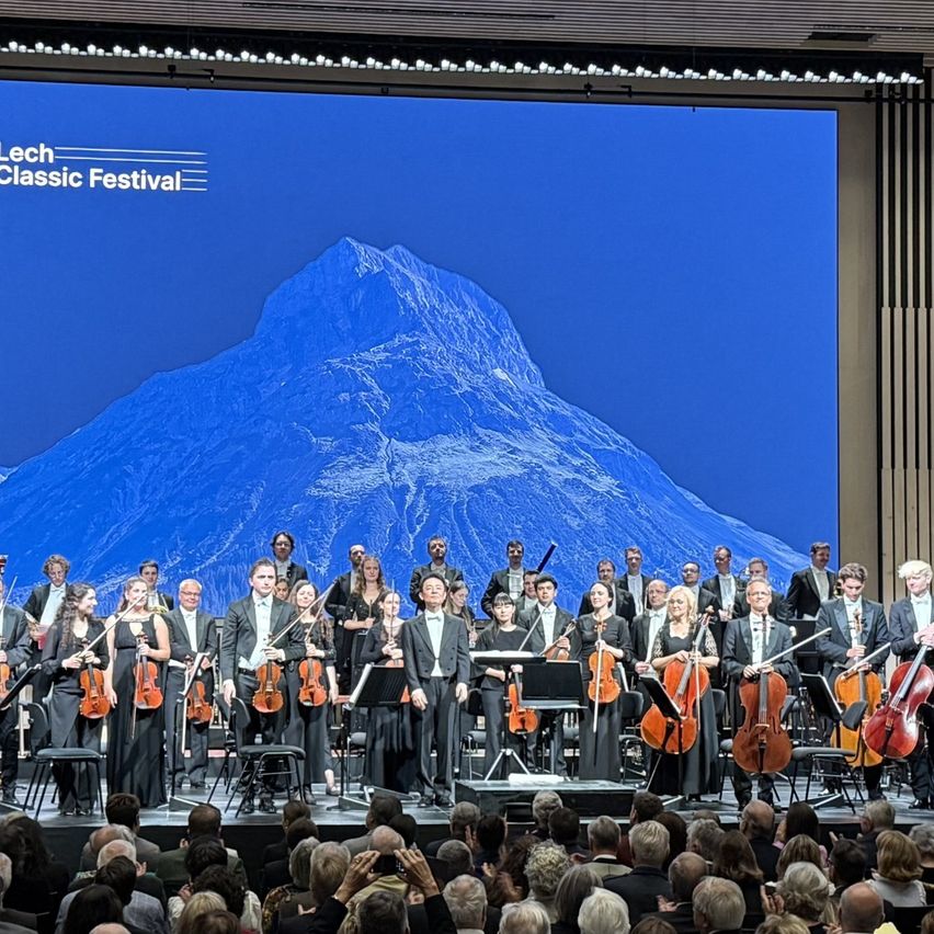 Ein großes Orchester spielt auf der Bühne vor einem Hintergrund eines Berges. Das Publikum sitzt vor ihnen.