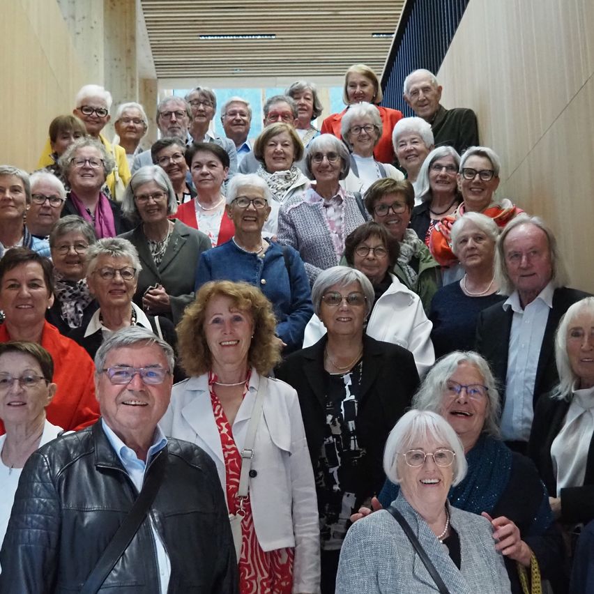 Eine große Gruppe von Menschen, meist ältere, lächeln und posieren für ein Gruppenfoto in einem Saal mit Holzwänden. Sie tragen verschiedene Outfits und Accessoires.