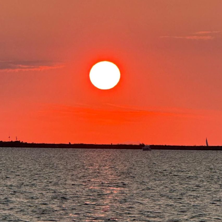 Die Sonne geht über einer ruhigen Wasserfläche unter, wobei ein kleines Boot am Horizont sichtbar ist. Der Himmel ist mit warmen, roten Farbtönen erfüllt.