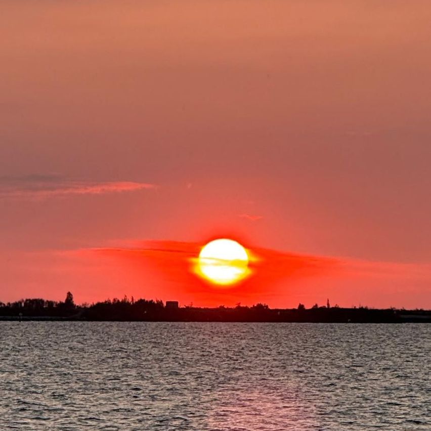 Die Sonne geht über einer ruhigen Wasserfläche unter und taucht den Himmel und die entfernten Bäume in ein warmes Licht.