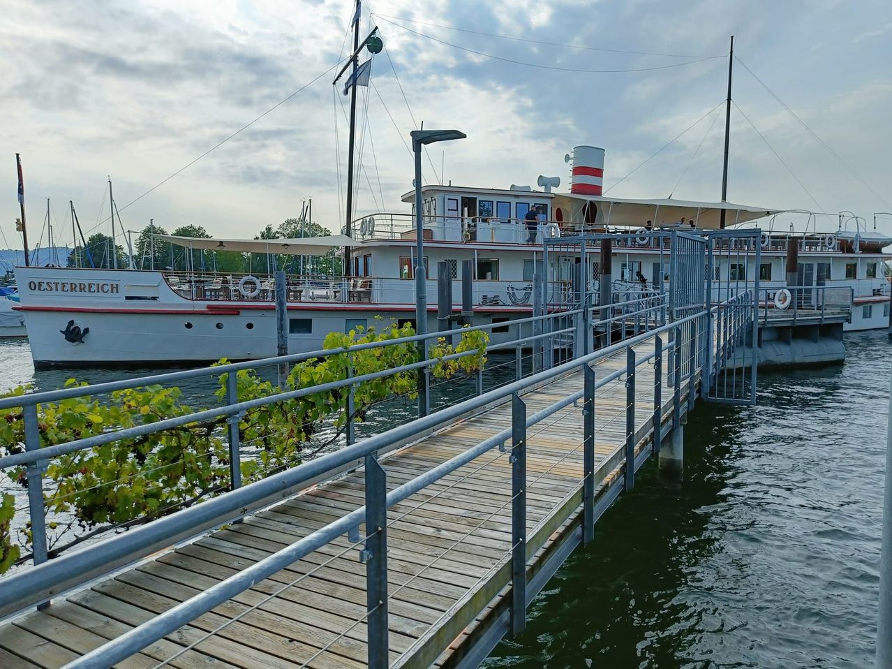 Eine Fähre liegt am Ufer eines Flusses, mit einer Holzbrücke, die zu ihr führt. Das Boot hat einen rot-weiß gestreiften Schornstein und eine weiße Markise.