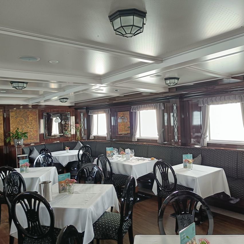 Ein leeres Restaurant mit Tischen und Stühlen, die mit weißen Tischdecken bedeckt sind. Der Raum hat einen Holzboden, weiße Decken und eine Bank mit gemusterter Rückenlehne. Es gibt mehrere Lampen und Fenster mit Vorhängen.