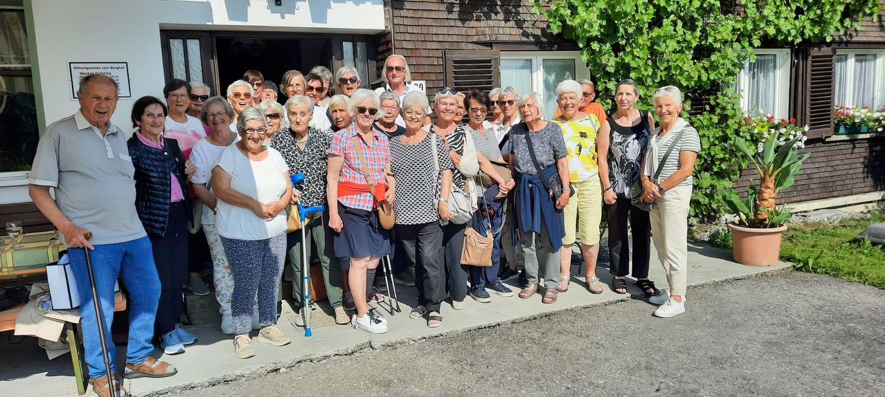 Eine Gruppe älterer Frauen, einige mit Gehstöcken, stehen auf einem Bürgersteig, lächeln und posieren für ein Foto vor einem Gebäude.