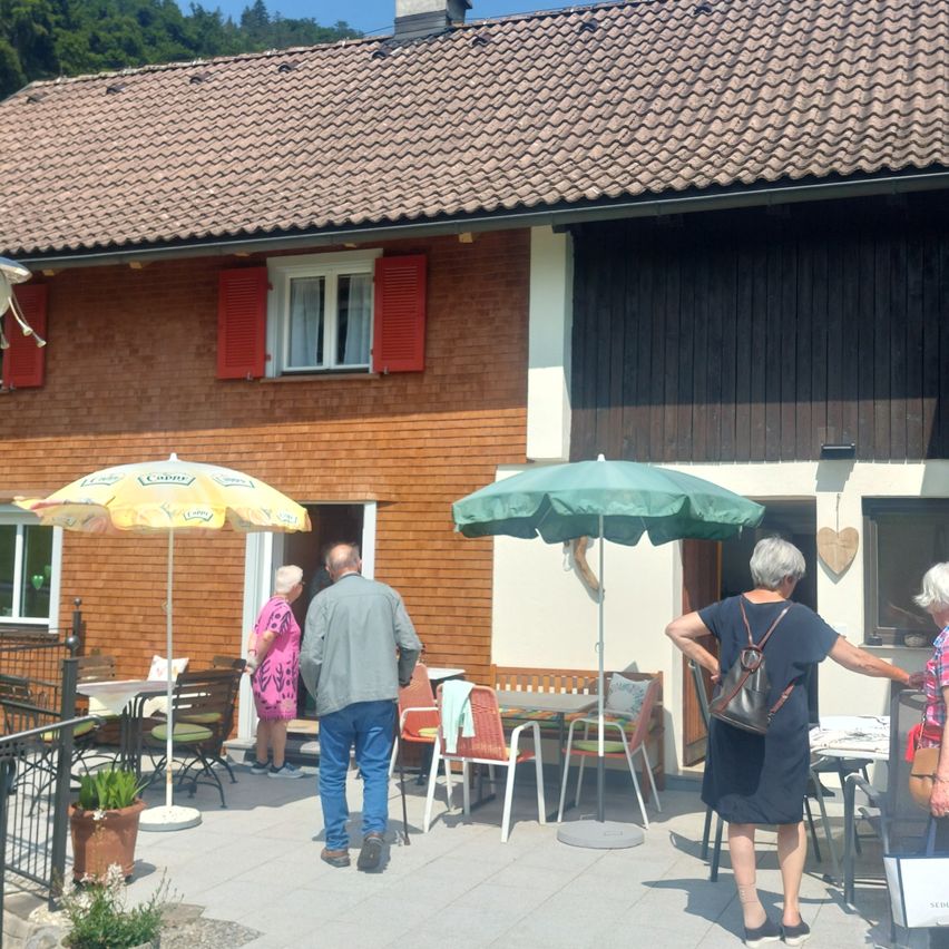 Eine Gruppe von Menschen steht vor einem Haus mit braunem Dach und roten Fensterläden. Zwei Frauen und ein Mann blicken zum Haus, während ein Regenschirm in der Nähe steht. Stühle und Tische sind draußen aufgestellt.