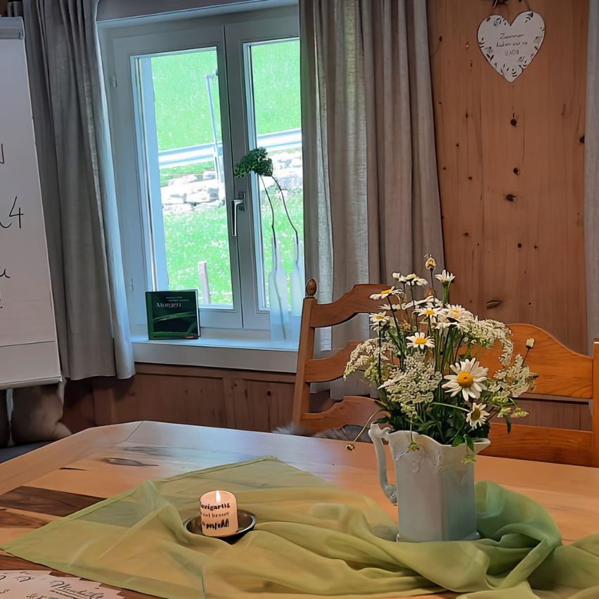 Ein Raum mit einer Holzwand und einem Fenster. Eine Kerze steht auf dem Tisch, und eine Vase mit Blumen steht auf einem Tisch. Ein Holzstuhl befindet sich rechts vom Tisch. Eine Tafel steht links.