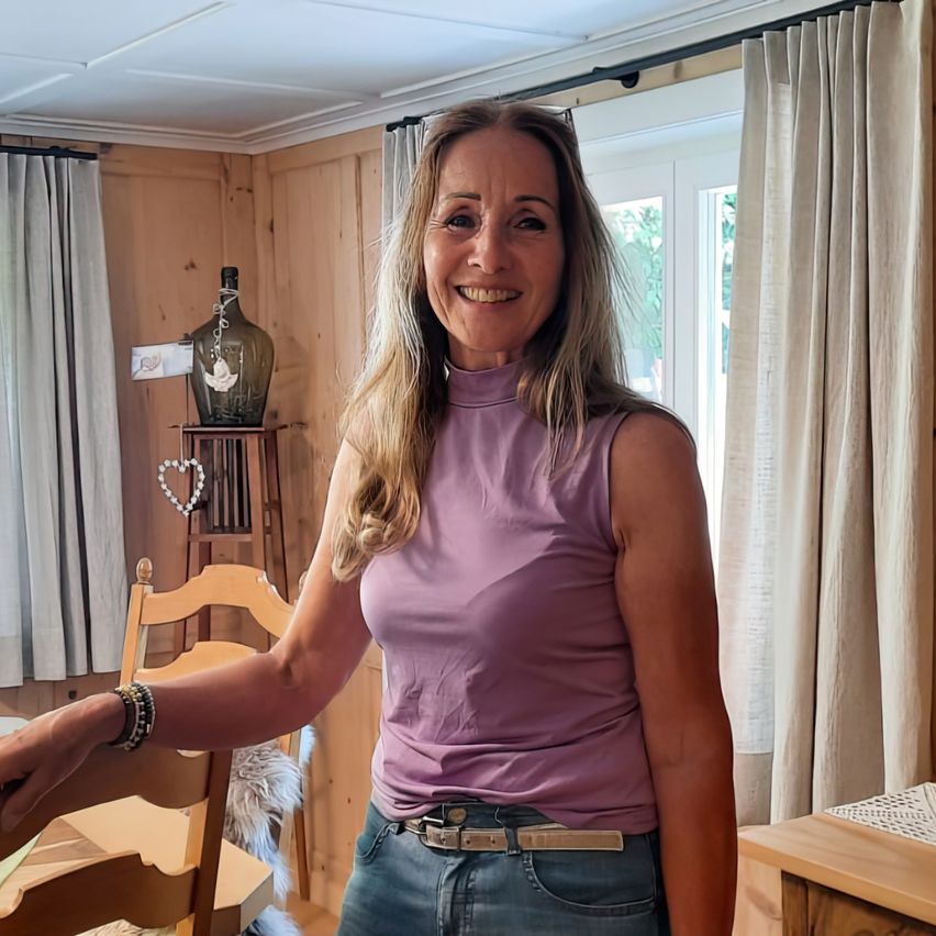 Eine lächelnde Frau mit blonden Haaren steht in einem gemütlichen Zimmer. Sie trägt ein lila ärmelloses Oberteil und Jeans. Hinter ihr befindet sich ein Holztisch mit Stühlen und ein dekoratives Element auf einem Regal.