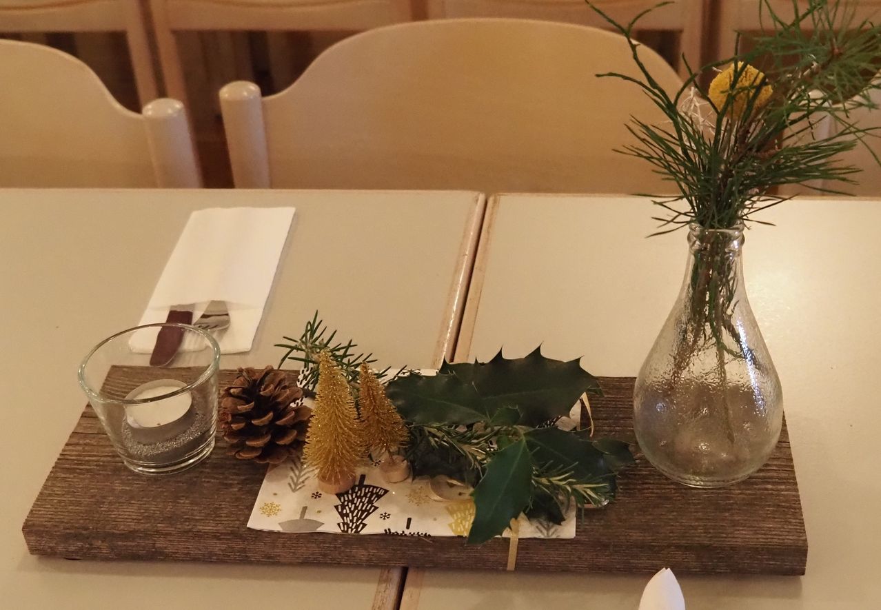 Eine Tischdekoration besteht aus einem Glas, einem Holztablett mit Tannenzapfen, kleinen Bäumen und Grünpflanzen sowie einer Vase mit Blumen. Links sind eine gefaltete Serviette, eine Gabel und ein Messer.