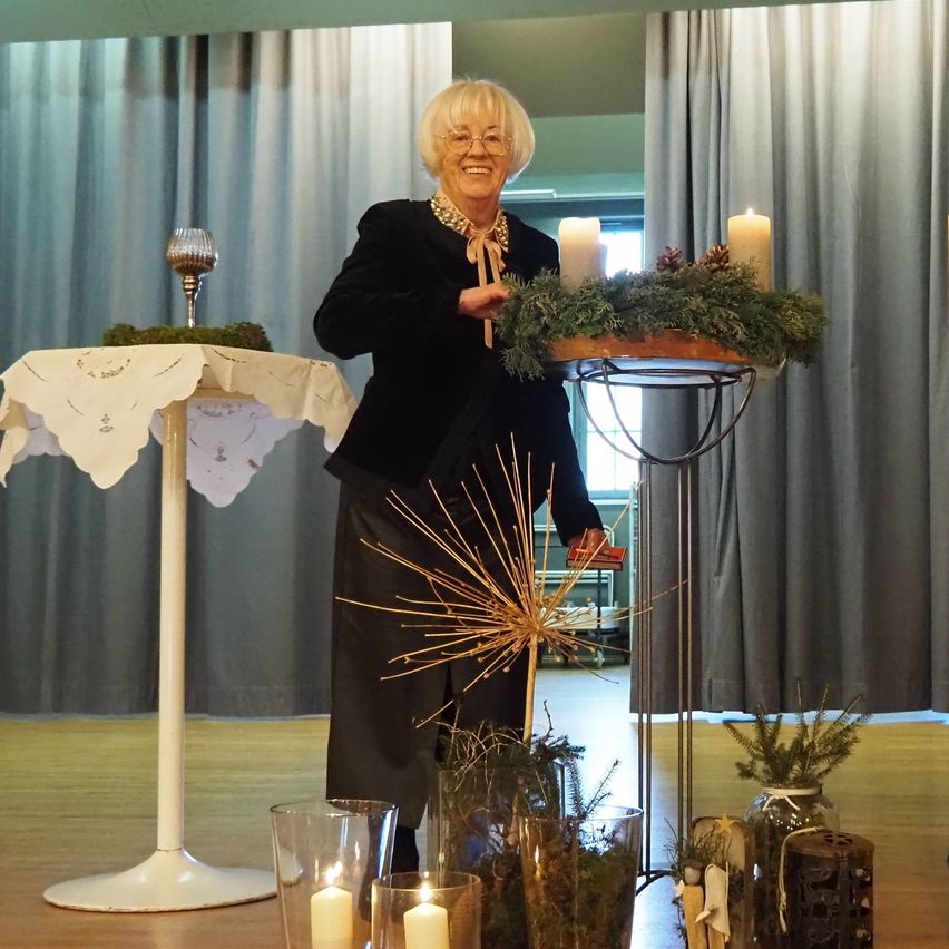 Eine ältere Frau mit Brille steht an einem Podium mit einem Blumenarrangement und zwei Kerzen und lächelt vor einem grauen Vorhang.