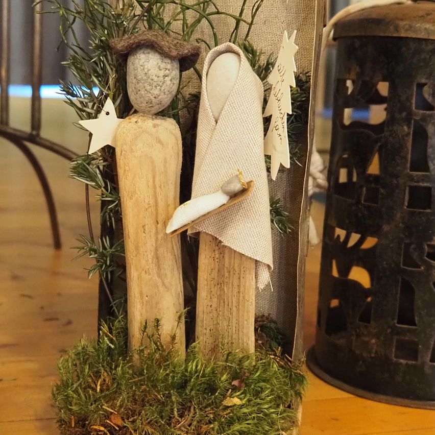 Zwei handgefertigte Figuren auf einer Holzbasis, eine trägt einen braunen Hut, die andere ist in weißes Gewebe gehüllt, umgeben von Grün und einem Weihnachtsbaum.
