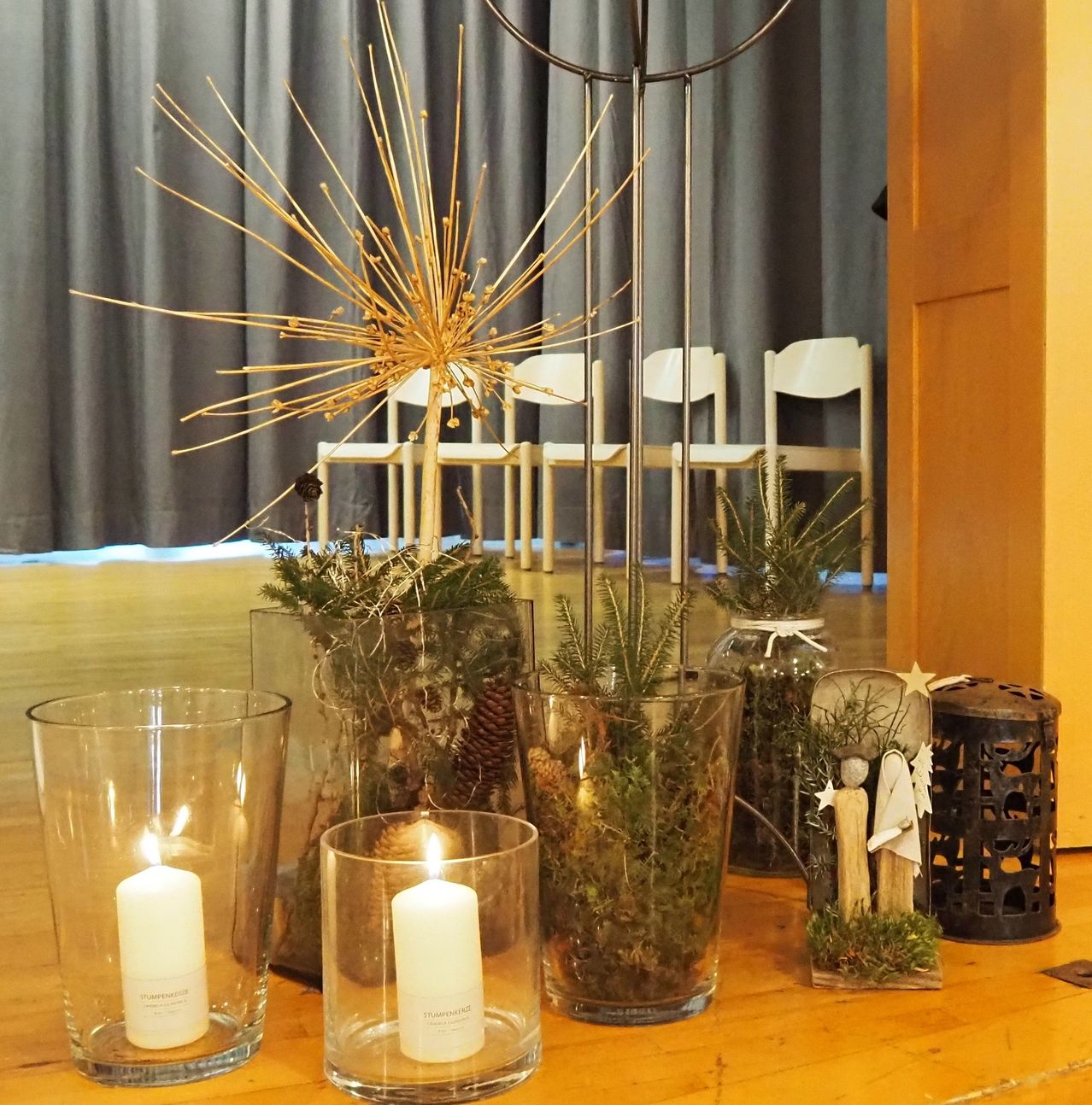 Ein Holztisch zeigt Kerzen und Pflanzen. Dahinter sind drei Stühle vor einem Vorhang aufgestellt. Eine goldene Struktur und Figuren sind im Vordergrund.