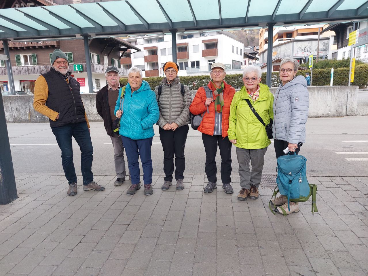 Eine Gruppe von sechs Personen, darunter drei Frauen und drei Männer, steht auf einem gepflasterten Gehweg. Sie sind in warme Kleidung gekleidet und halten Rucksäcke. Sie sind wahrscheinlich Touristen. Hinter ihnen befindet sich eine Überdachung mit Glasscheiben.