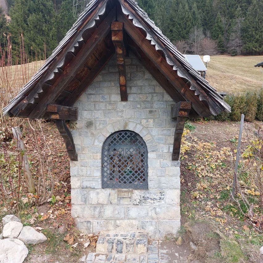 Eine kleine Kapelle mit einer Steinmauer und einem Holzdach befindet sich in einem Grasbereich. Die Kapelle hat ein Fenster mit einem Metallgitter. Im Hintergrund befinden sich Bäume und Büsche.