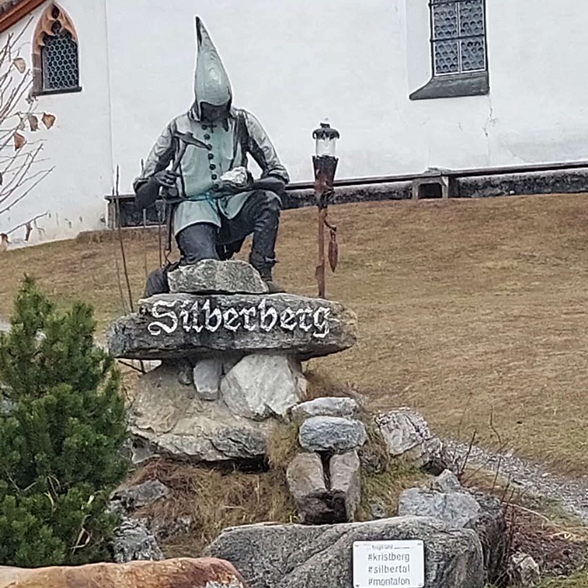 Eine Statue einer Figur mit Spitzhut und Hammer sitzt auf einem Felsen mit der Aufschrift Silberberg vor einem Gebäude mit Bogenfenstern.