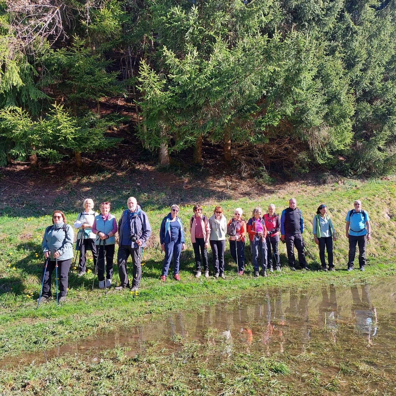 A group of hikers poses for a photo in a grassy field with trees in the background. They are all dressed for outdoor activities, some holding trekking poles.