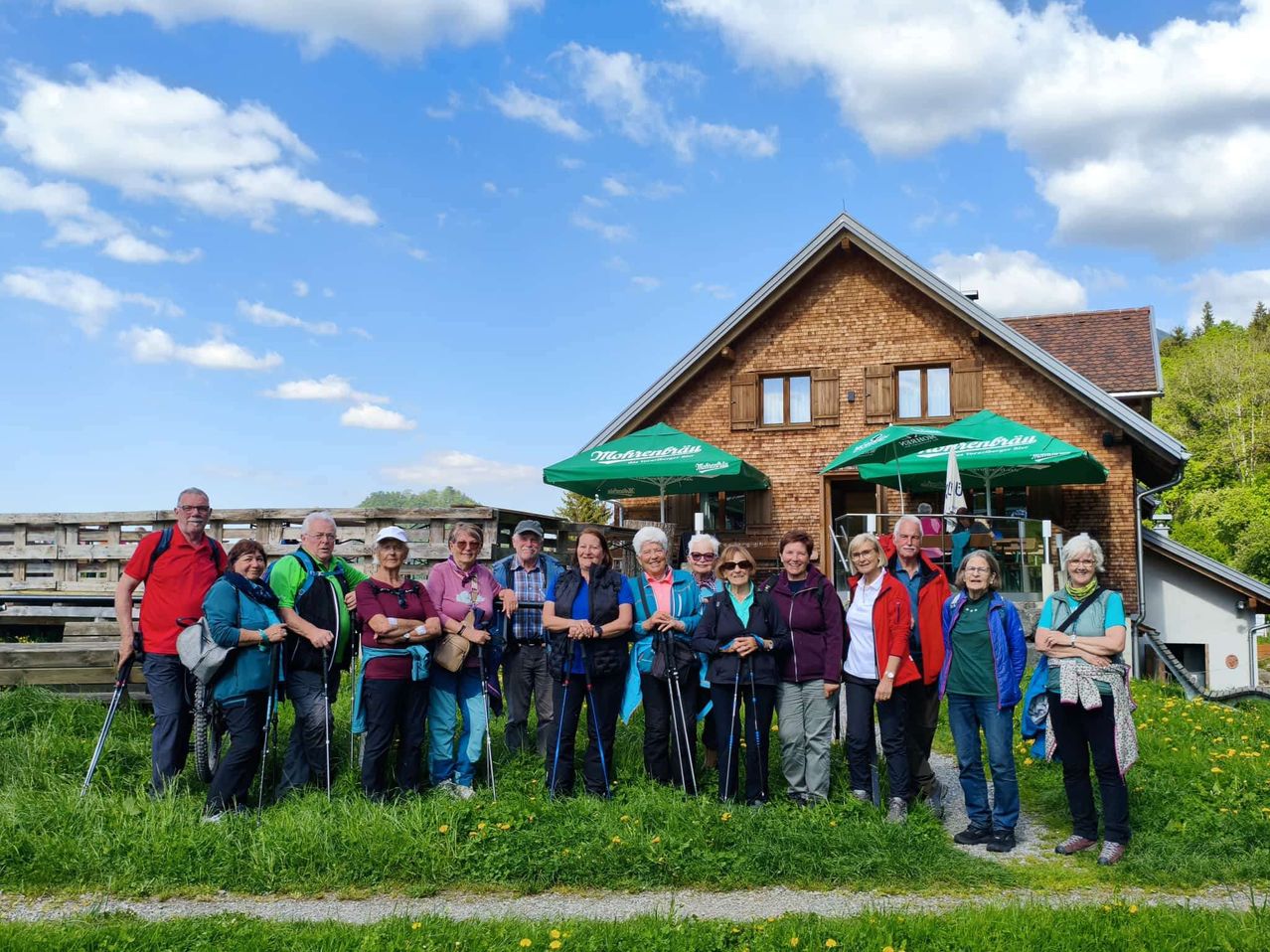Eine Gruppe von Menschen, die zum Wandern gekleidet sind, steht vor einem Backsteinhaus mit grünen Schirmen. Sie posieren für ein Foto an einem sonnigen Tag.