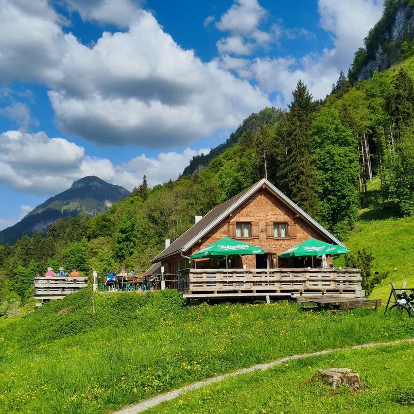 Ein Bergchalet mit grünen Schirmen auf einem grasbewachsenen Hang unter einem blauen Himmel mit Wolken. Leute sitzen draußen und genießen die Aussicht.