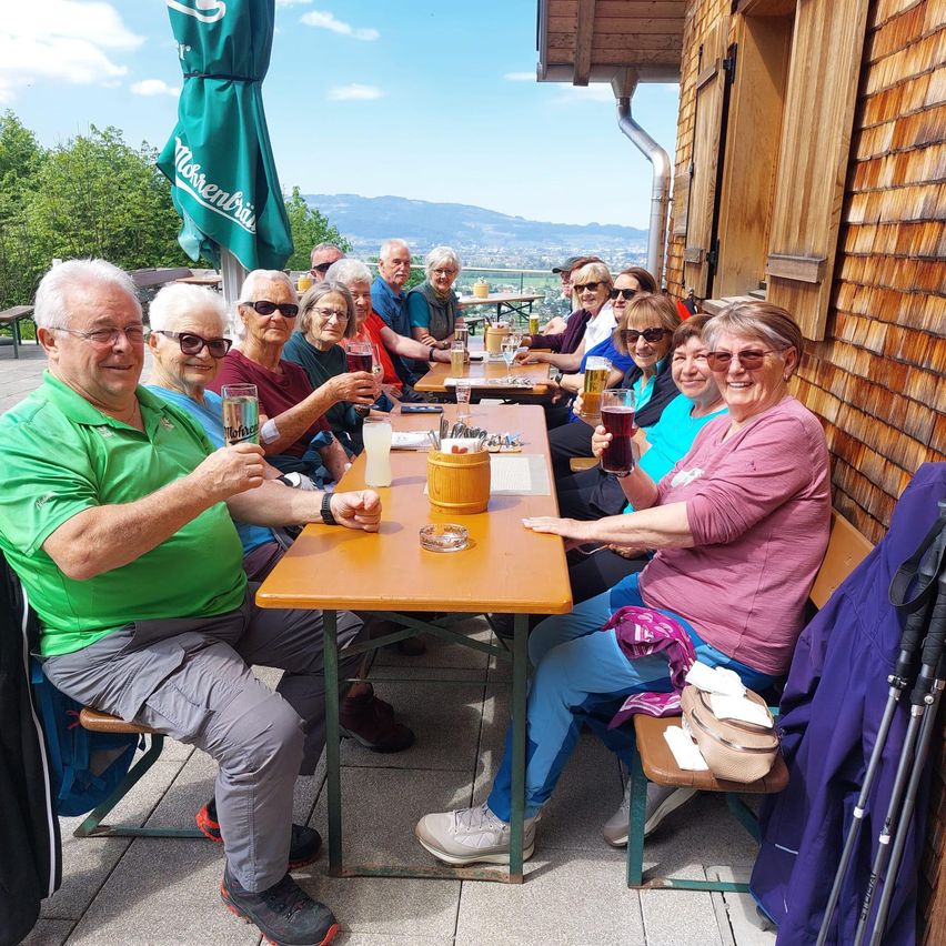 Eine Gruppe von Menschen sitzt an einem Tisch im Freien und hält Gläser mit Bier in den Händen. Sie lächeln und scheinen ihre Mahlzeit zu genießen. Der Tisch ist mit Tassen und Tellern gedeckt.