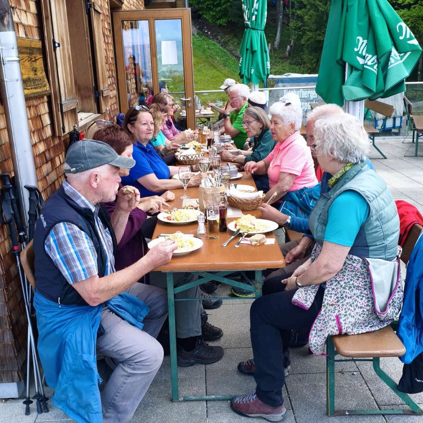 Eine Gruppe älterer Personen sitzt um einen Tisch in einem Außenbereich. Sie genießen ihr Essen mit Tellern, Getränken und Besteck. Im Hintergrund befinden sich Regenschirme und eine Holzwand.