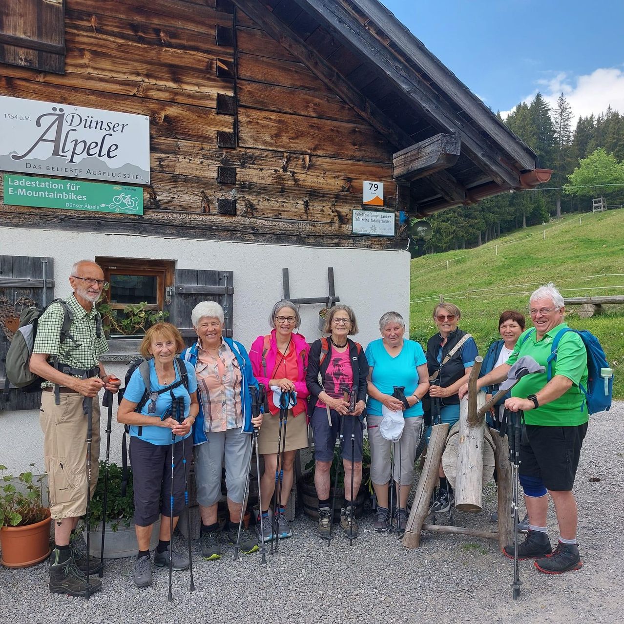 Eine Gruppe von Wanderern posiert für ein Foto vor einer Holzchalet mit der Aufschrift 'Dünser Alpele', mit einem grünen Schild für E-Mountainbikes. Sie halten Trekkingstäbe und Rucksäcke.