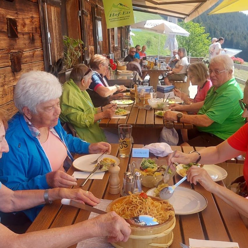 Eine Gruppe älterer Erwachsener isst in einem Restaurant. Sie sitzen um einen Holztisch herum. Die Frau in der blauen Jacke schneidet ein Gericht.