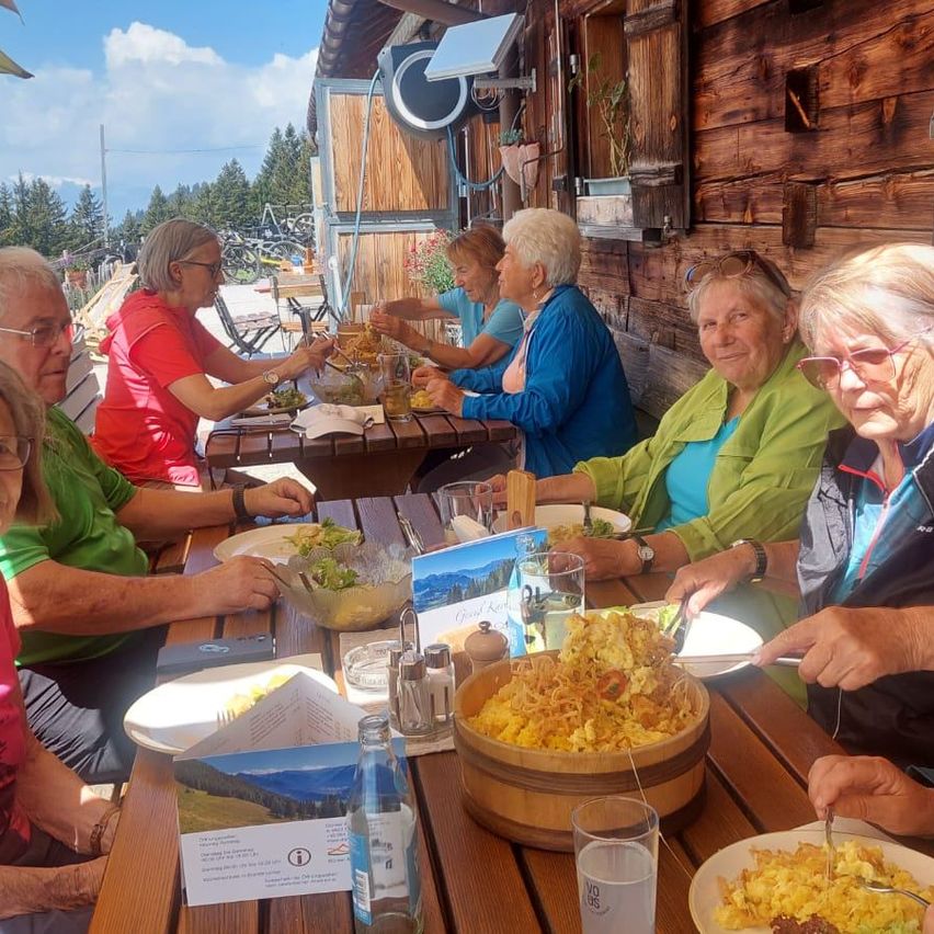 Mehrere Frauen sitzen an einem Holztisch in einem Outdoor-Restaurant. Sie essen verschiedene Gerichte, darunter Salate und Pasta. Der Tisch ist mit Tellern, Gläsern und Gewürzen bestückt. Dahinter befindet sich eine Holzwand mit Pflanzen und Fahrrädern.
