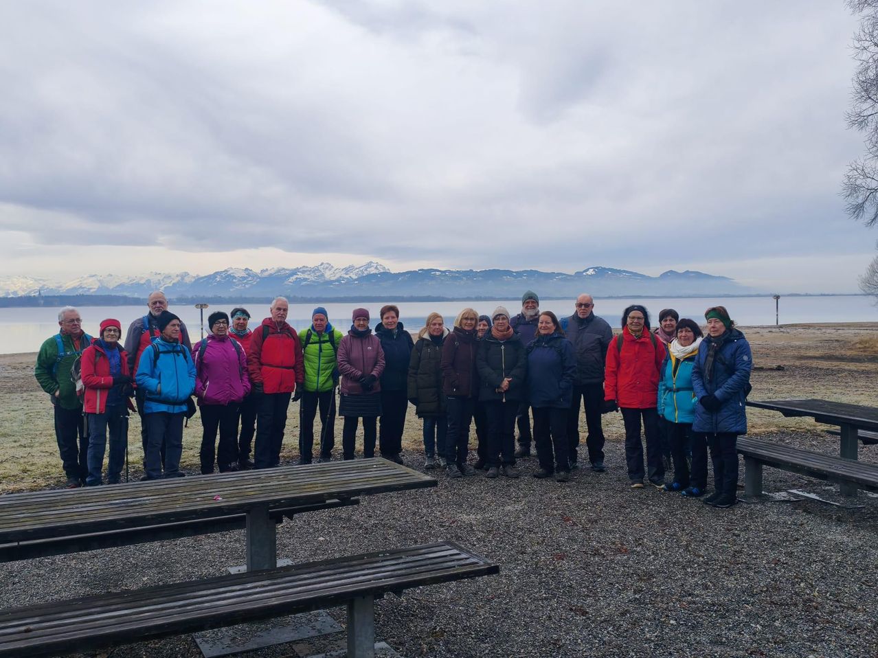 A group of people stands in front of a lake with mountains in the background, dressed in warm clothing. They are smiling and appear to be posing for a photo.