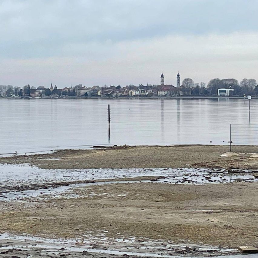 A wide river landscape with a city skyline in the distance, featuring two prominent towers and several buildings under a cloudy sky.