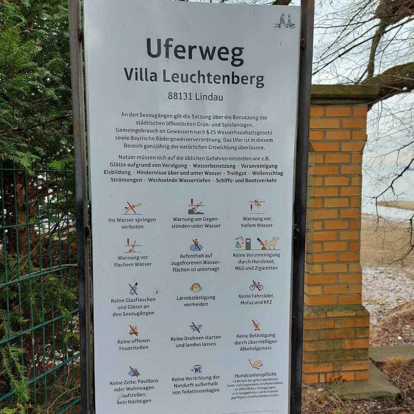 A sign by a lake reads Uferweg Villa Leuchtenberg 88131 Lindau. It includes regulations for using the lakeside, such as prohibiting water jumping, drinking water, and littering. It also warns against swimming under water, feeding animals, and using bicycles, motorbikes, and cars.