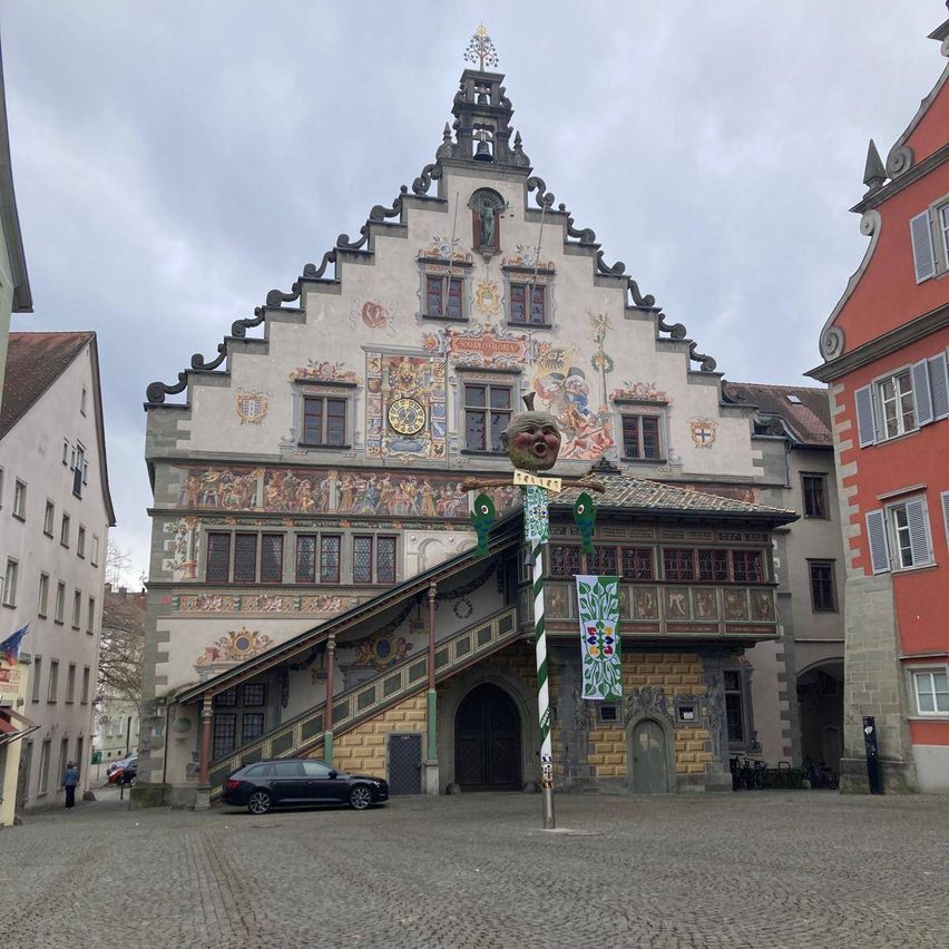 A historic building with intricate designs and a clock tower stands in a town square. A black car is parked in front, and people walk nearby.