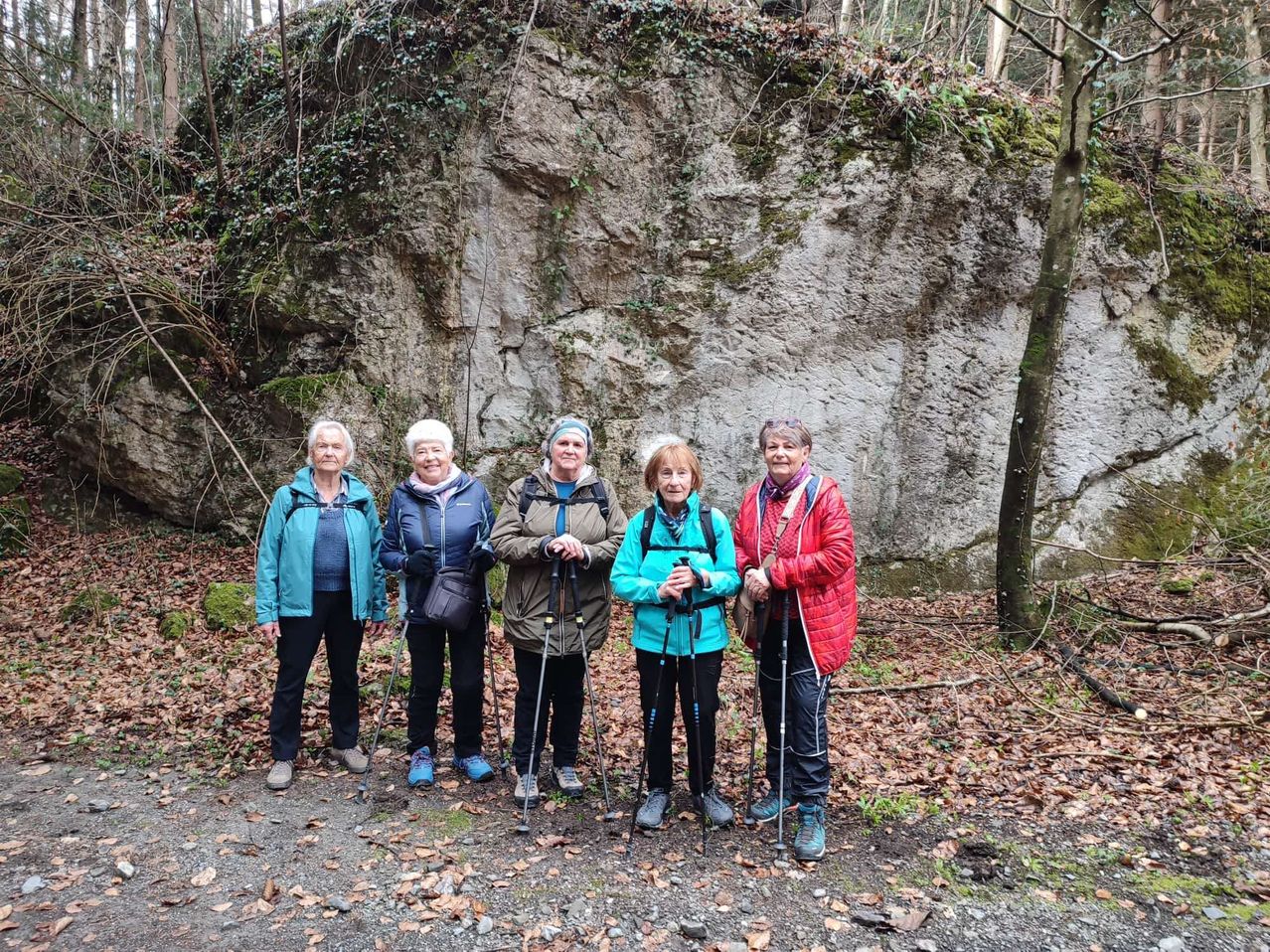 Fünf Frauen stehen in einem Wald und tragen alle Wanderausrüstung. Sie posieren für ein Foto mit einer großen Felswand im Hintergrund.