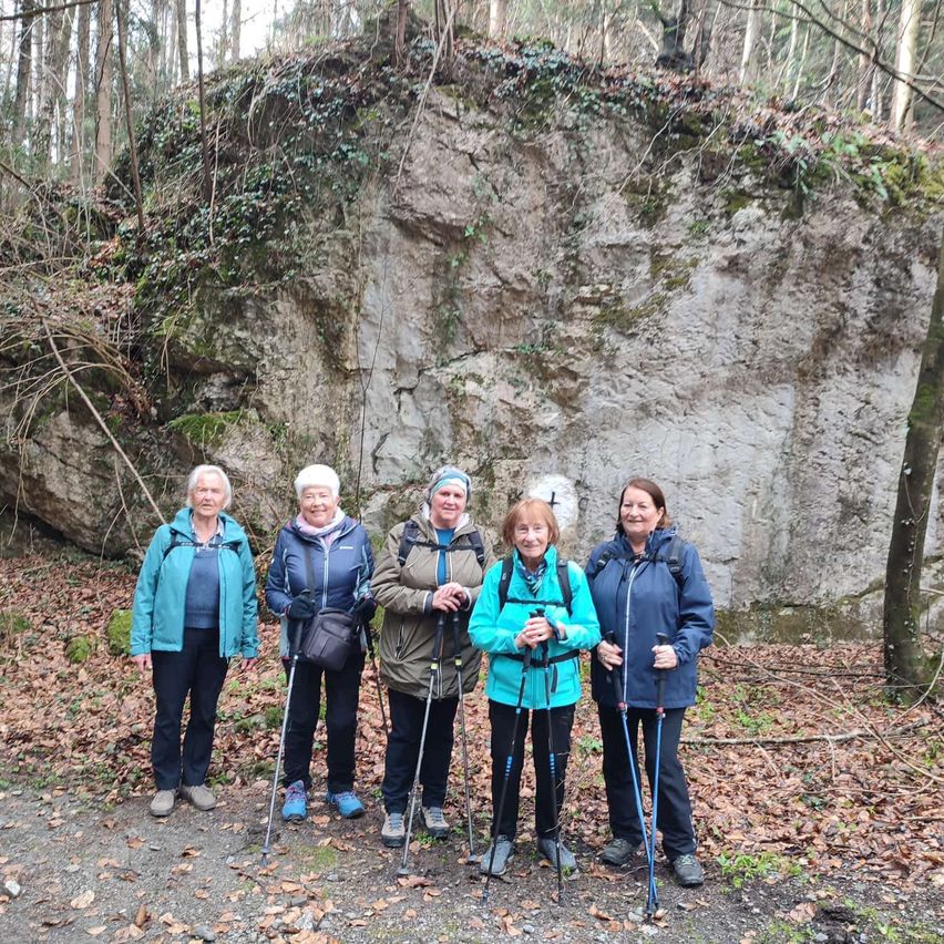 Fünf Frauen stehen in einem Wald, alle tragen Jacken und halten Wanderstöcke. Sie posieren vor einem großen Felsen.