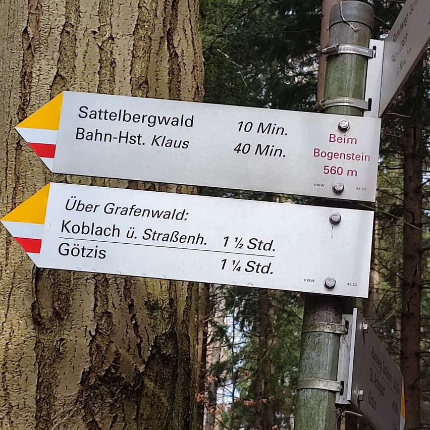 Ein Wegweiser in der Nähe eines Baumes zeigt Wanderrouten an. Er weist 10 Minuten zu Sattelbergwald und 40 Minuten zu Bahn-Hst. Klaus aus. Ein weiterer Schild zeigt Routen über Grafenwald.