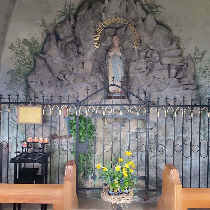 Eine kleine Kapelle zeigt eine Statue der Jungfrau Maria mit einem steinernen Hintergrund. Die Kapelle ist von einem schwarzen Metalltor umgeben und hat einen Tisch mit brennenden Kerzen. Vor dem Tor steht ein Blumenarrangement in einem geflochtenen Korb.