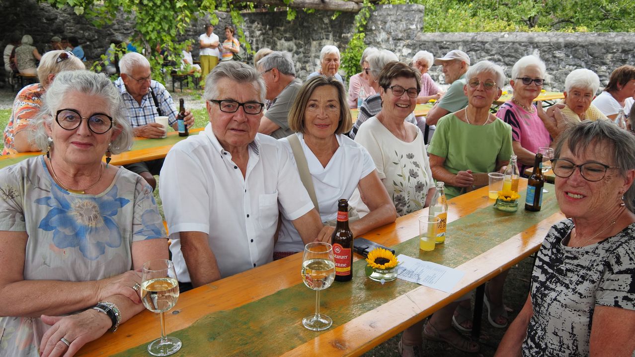 Eine Gruppe älterer Menschen sitzt draußen an einem Tisch und genießt Getränke und Essen. Der Tisch ist mit Weingläsern, einer Flasche und einer Sonnenblume gedeckt.