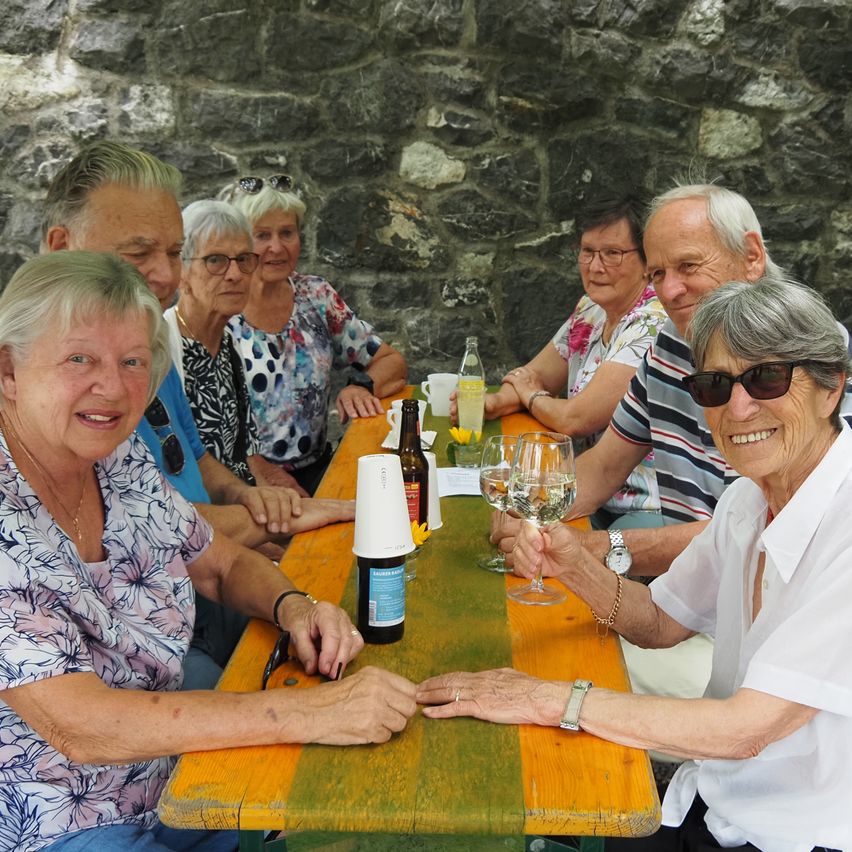 Eine Gruppe älterer Menschen sitzt um einen Tisch herum, lächelt und hält Weingläser in der Hand. Sie befinden sich in einer Außenumgebung mit einer Steinmauer im Hintergrund.