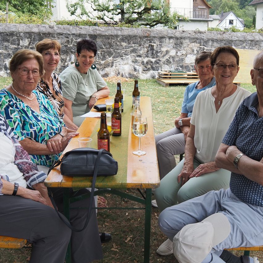Eine Gruppe älterer Frauen und Männer sitzt um einen Tisch in einem Garten, lächelt und posiert für ein Foto. Flaschen Bier und ein Glas Wein stehen auf dem Tisch. Dahinter befindet sich eine Steinmauer mit einem Haus und Bäumen.