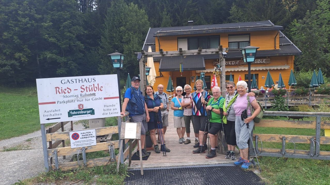A group of elderly hikers poses for a photo in front of a yellow building with a sign that reads 'Stahaus-Stihle'. They are holding trekking poles and wearing hiking gear. Behind them, there is a sign prohibiting parking.