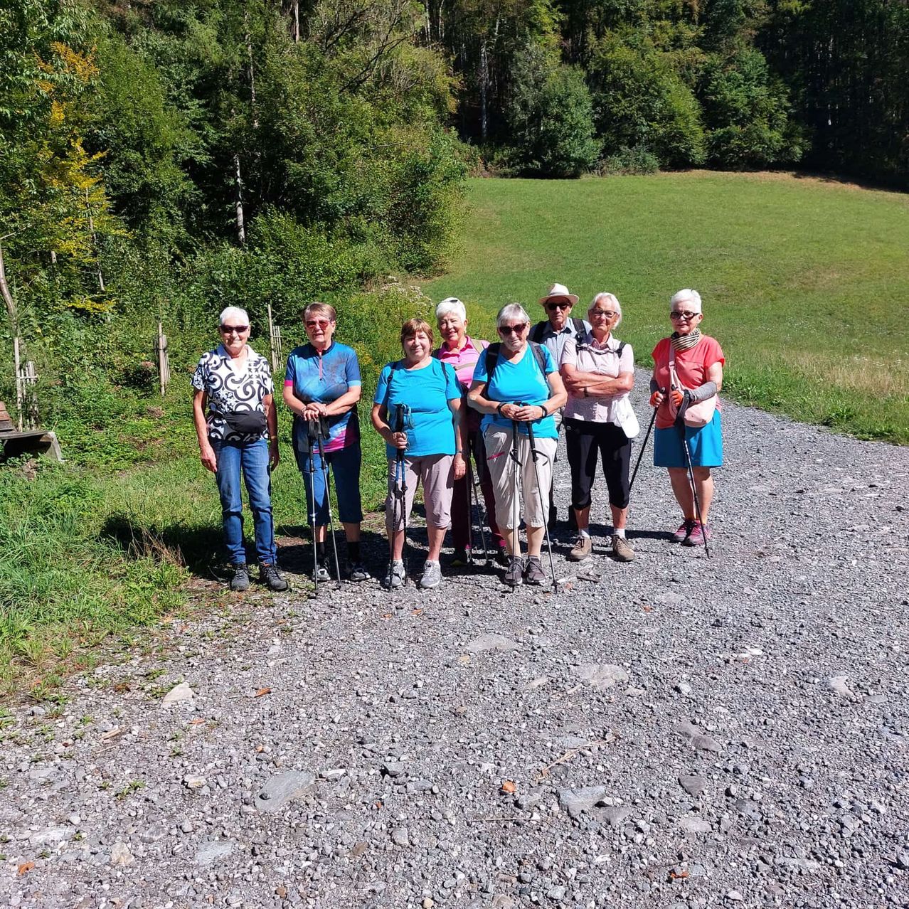 Eine Gruppe von Wanderfrauen steht auf einem Schotterweg, lächelt und posiert für ein Foto. Sie halten Trekkingstöcke und tragen Sonnenbrillen. Hinter ihnen befindet sich eine Wiese mit Bäumen.