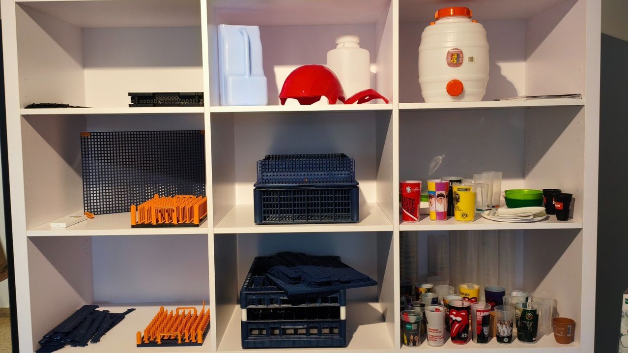 Ein Regal mit verschiedenen Gegenständen, darunter blaue und orangefarbene Kisten, ein roter Helm und ein weißer Kunststoffbehälter mit orangefarbenem Deckel.