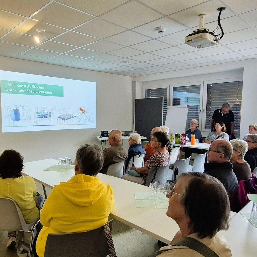 Eine Gruppe von Menschen sitzt in einem Raum und nimmt an einer Präsentation teil. Eine Projektionsleinwand zeigt Informationen an, und ein Mann steht in der Nähe einer Tafel. Der Raum ist gut beleuchtet, und es gibt Tische mit Gläsern und anderen Gegenständen.