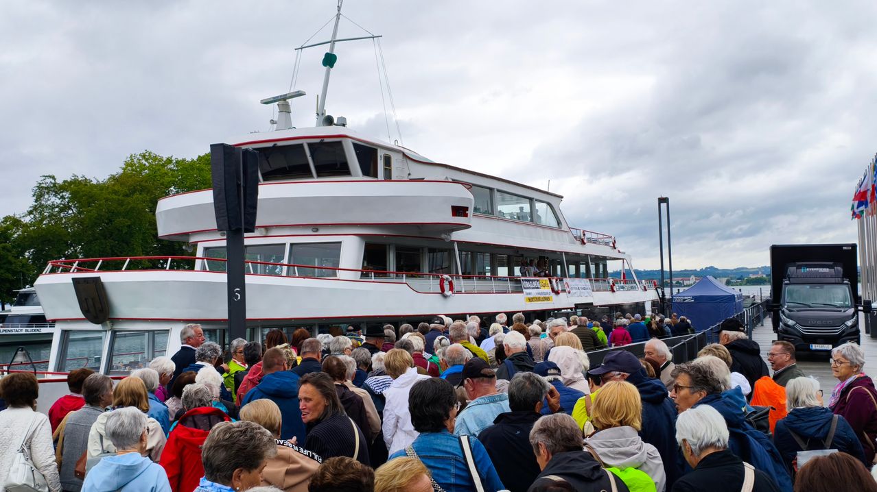 A crowd of people gathers around a large ferry boat with a white and red hull, under a cloudy sky. Many individuals wear jackets and hats, and some carry bags.