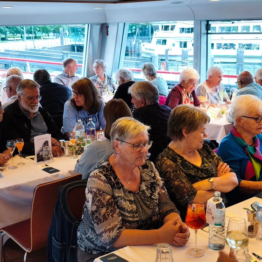 Several elderly people are sitting in a restaurant with large windows, enjoying drinks and conversation. Outside, there are boats and a train track visible.