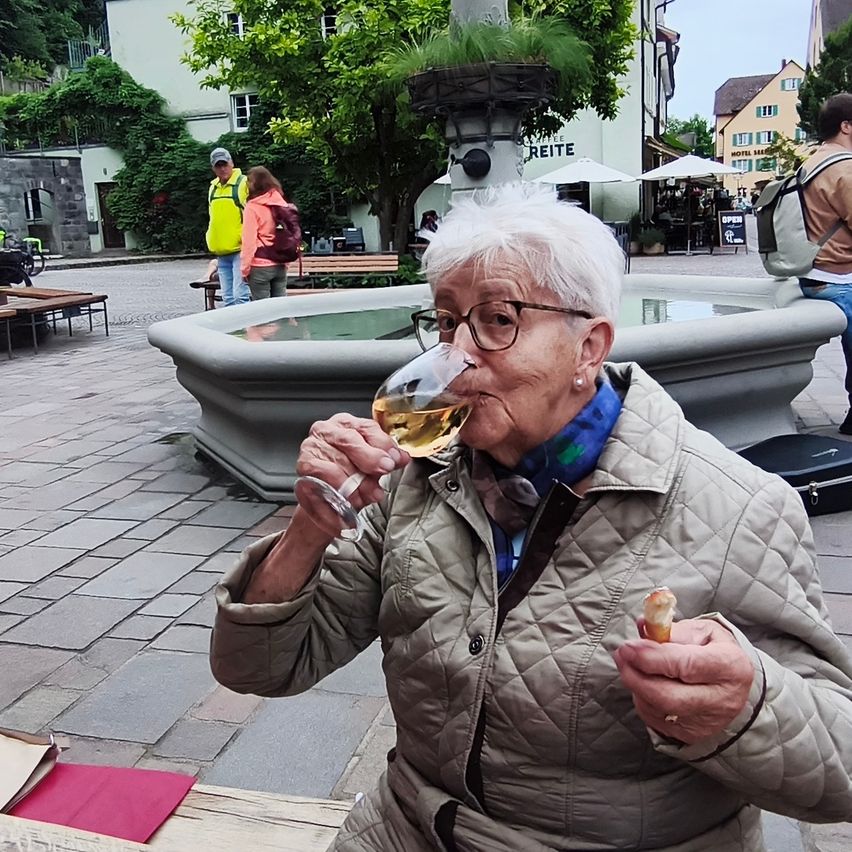 An elderly woman with glasses is drinking wine at a fountain, seated on a bench with a small pastry in her hand. Behind her are two people, a man with a backpack, and a woman with a backpack.