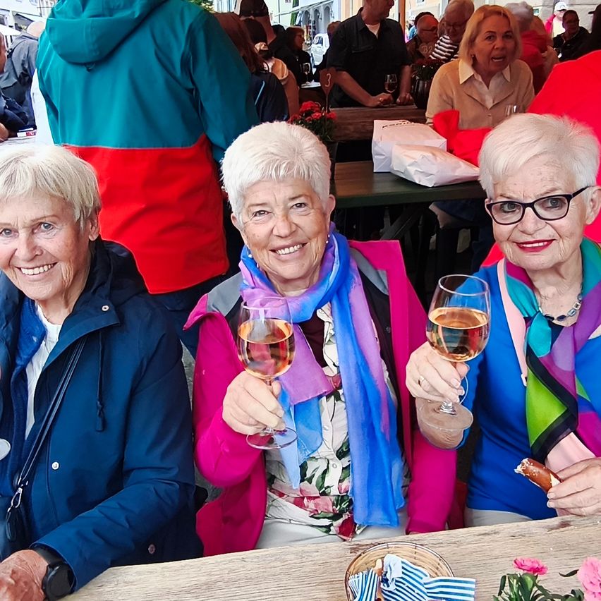 Three elderly women, all smiling, pose for a photo at an outdoor event. One holds a glass of wine, another has a sausage, and they wear colorful attire. Blurred figures of people and tables are in the background.