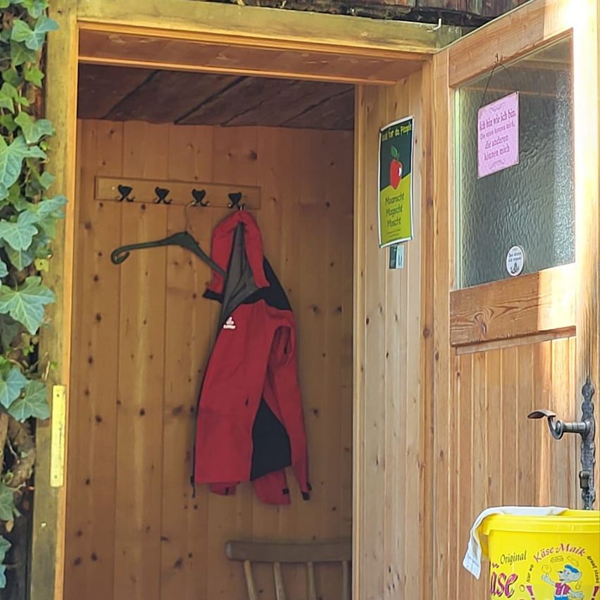 Ein Holzschuppen mit offener Tür, auf der ein roter Mantel an einem Haken hängt, ein Holzstuhl und ein Schild mit einem Apfel darauf.